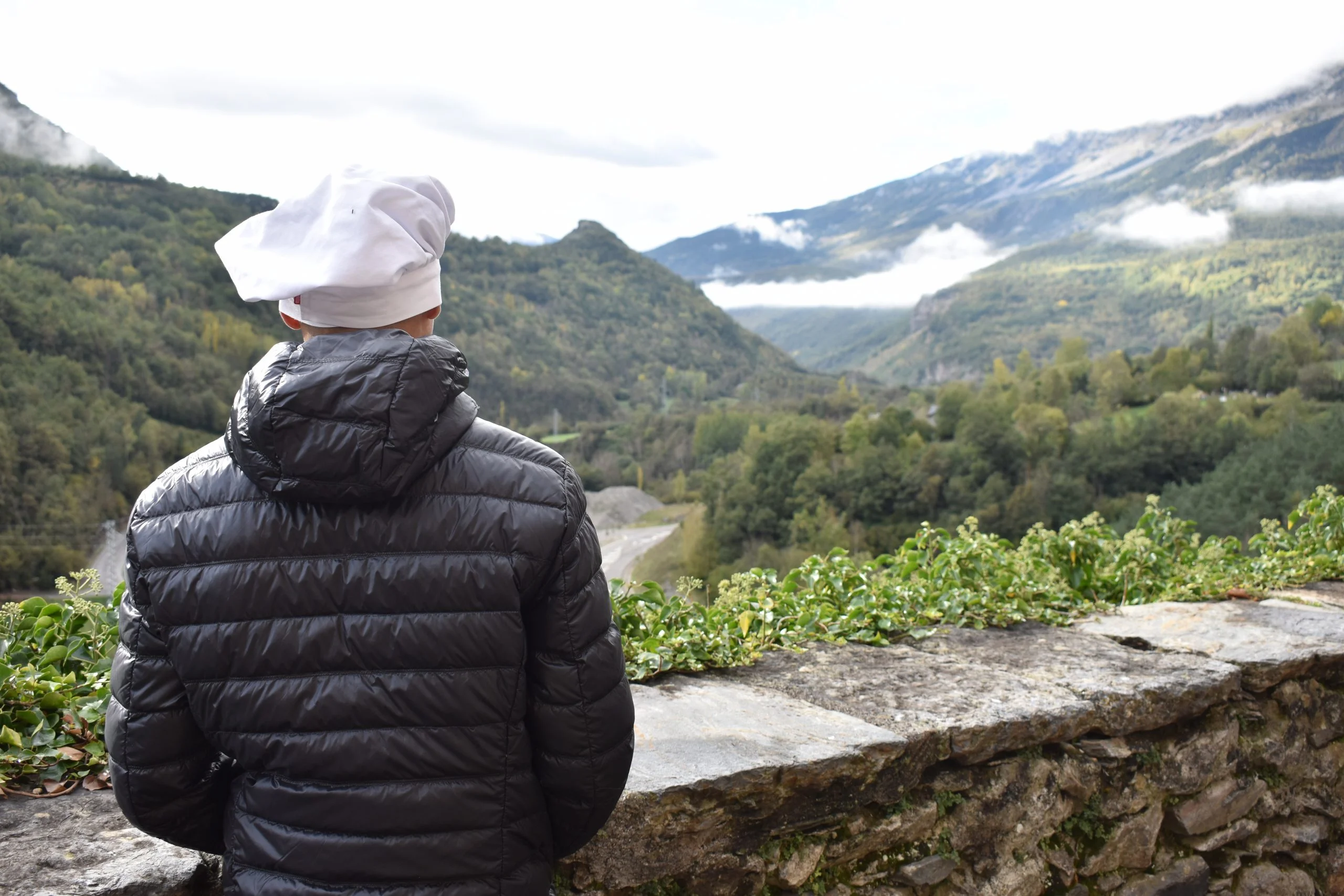 Un alumno de la escuela de hosteleria guayente, con abrigo y gorro de chef, observa el horizonte desde la terraza de la entrada del Santuario de Guayente. Al fondo se ve la ladera de la Sierra de Chía con los bosques.